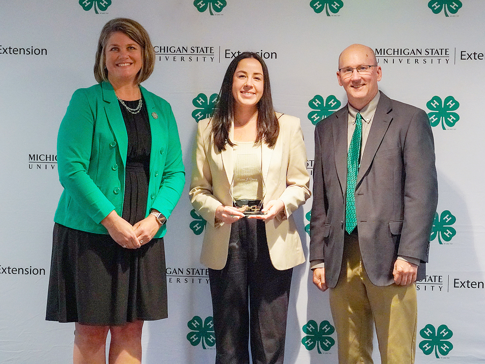 Three individuals at a backdrop. The center person holds an award.
